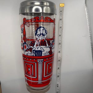 Pode incluir: Coqueteleira vintage com tampa de a&ccedil;o inoxid&aacute;vel. O corpo de vidro transparente apresenta uma ilustra&ccedil;&atilde;o vermelha e branca de um homem de bigode atr&aacute;s de um bar, com a palavra "BEER" vis&iacute;vel. A coqueteleira tem aproximadamente 23 cm de altura.