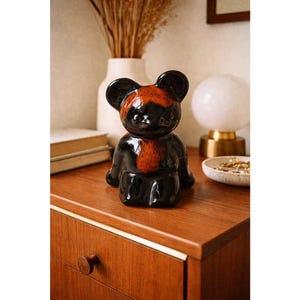May include: A ceramic bear figurine with a glossy black finish and a reddish-brown chest and head. The bear is sitting on a wooden surface, with a white vase and a lamp in the background. The figurine is a decorative item.