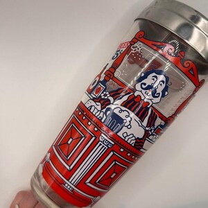 Pode incluir: Uma coqueteleira de vidro vintage com tampa de a&ccedil;o inoxid&aacute;vel. A coqueteleira apresenta um design vermelho, branco e azul com um personagem de desenho animado segurando uma cerveja. O design inclui a palavra "Beer".