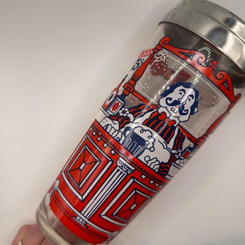 Pode incluir: Uma coqueteleira de vidro vintage com tampa de a&ccedil;o inoxid&aacute;vel. A coqueteleira apresenta um design vermelho, branco e azul com um personagem de desenho animado segurando uma cerveja. O design inclui a palavra "Beer".