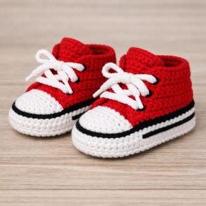 Op de afbeelding: Een paar handgemaakte, gehaakte babysneakers in rood en wit. De sneakers hebben witte veters, een witte neus en een zwarte streep langs de zool. Ze worden gepresenteerd op een licht houten oppervlak.