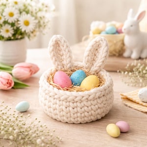 Puede incluir: Una cesta de punto color crema con forma de conejo, que contiene tres huevos de Pascua pastel. La cesta tiene dos orejas verticales y está llena de paja. Huevos adicionales y un conejo de cerámica están en el fondo, sugiriendo un tema de Pascua.