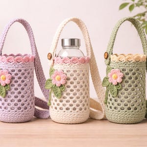 Op de afbeelding: Drie gehaakte waterfles houders in lavendel, crème en saliegroen. Elk heeft een bloemversiering en een riem. De houders zijn versierd met een geschulpte rand en een knoopsluiting. De houders zijn ontworpen om een waterfles te dragen.