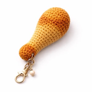 May include: A crocheted chicken leg keychain in shades of orange and yellow. The keychain has a gold-colored clasp and a small, round, off-white bead charm. The chicken leg is the main focus, set against a white background.