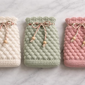 May include: Three handmade crochet pouches in cream, sage green, and pink. Each pouch has a textured, diagonal stitch pattern and a scalloped top. They are tied with a decorative string and wooden beads. The pouches are likely for small items.