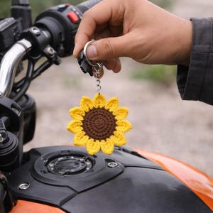 Puede incluir: Un llavero de girasol de ganchillo amarillo y marrón se muestra frente a una motocicleta negra y naranja. El llavero está hecho a mano y está sujeto a un llavero. Se ven los manillares de la motocicleta.