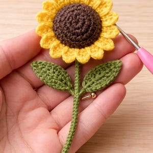 May include: A handmade crochet sunflower with yellow petals and a brown center. The flower has a green stem and leaves, and a gold-colored pin. The craft project is held in a hand.