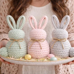 Puede incluir: Tres figuras de conejitos de Pascua de ganchillo en colores pastel: verde menta, rosa y gris. Cada conejito tiene una cola esponjosa y un lazo de cuerda. Están sobre un plato de cerámica moteada con huevos de Pascua decorativos y paja.