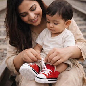 Op de afbeelding: Een persoon bindt de veters van rode en witte gehaakte babyschoenen. De schoenen hebben een zwarte streep en witte veters. De baby draagt een wit t-shirt en een kaki korte broek. De persoon draagt een beige outfit.