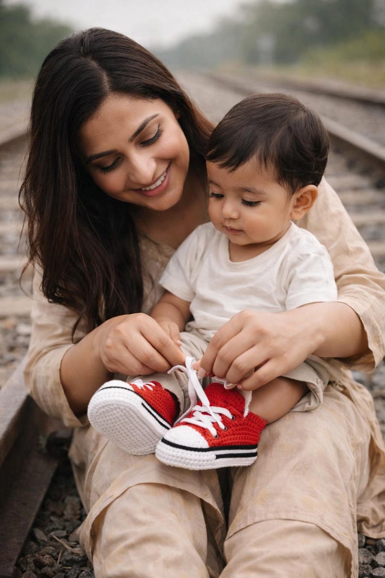 Op de afbeelding: Een persoon bindt de veters van rode en witte gehaakte babyschoenen. De schoenen hebben een zwarte streep en witte veters. De baby draagt een wit t-shirt en een kaki korte broek. De persoon draagt een beige outfit.