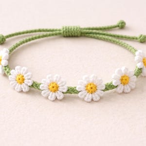 Könnte beinhalten: Ein handgefertigtes Gänseblümchen-Armband mit weißen Häkelblumen mit gelben Zentren, auf einer grünen Kordel aufgereiht. Das verstellbare Armband ist ein bezauberndes Accessoire, perfekt für einen Hauch von Natur.