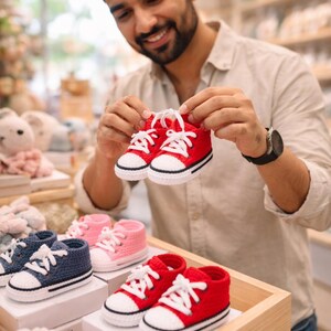 Op de afbeelding: Een paar rode gehaakte babysneakers met witte veters. De sneakers hebben een zwarte streep langs de zool. Andere paren gehaakte babyschoenen in roze en marineblauw zijn ook zichtbaar.
