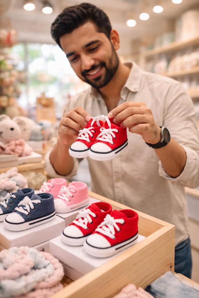 Op de afbeelding: Een paar rode gehaakte babysneakers met witte veters. De sneakers hebben een zwarte streep langs de zool. Andere paren gehaakte babyschoenen in roze en marineblauw zijn ook zichtbaar.