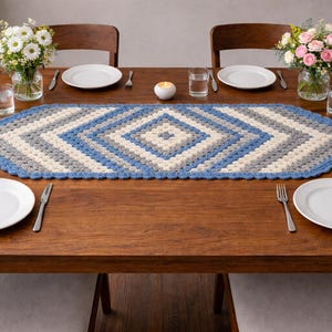 May include: A wooden dining table set for four with white plates, silverware, and glasses. A handmade, blue, gray, and white crochet table runner is centered on the table. Vases of flowers and a candle complete the setting.