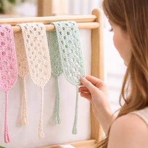 Puede incluir: Cuatro marcapáginas de ganchillo hechos a mano en colores pastel: rosa, crema y verde menta. Los marcapáginas se exhiben en un estante de madera. Cada marcapáginas tiene una borla larga y colgante. El diseño de tejido abierto es visible.
