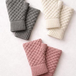 May include: Three pairs of fingerless gloves in different colors: gray, cream, and pink. Each pair features a ribbed cuff and a textured, diagonal stitch pattern. The gloves are made of a soft, knitted material.