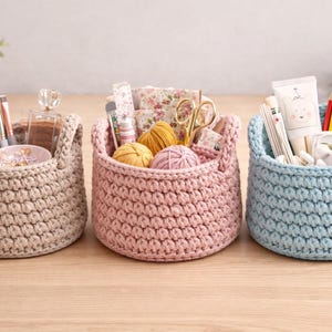 Puede incluir: Tres cestas de ganchillo hechas a mano en beige, rosa y azul. La cesta beige contiene brochas de maquillaje y perfume. La cesta rosa contiene lana y tijeras. La cesta azul está llena de lápices y un tubo de loción. Cada cesta tiene un asa.