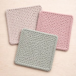 Può includere: Tre quadrati all'uncinetto fatti a mano in morbidi colori pastello: rosa chiaro, verde salvia e beige neutro. I quadrati sono disposti su una superficie di legno chiaro, mettendo in risalto la trama del punto all'uncinetto.