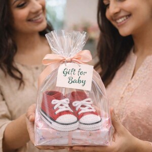 Op de afbeelding: Een paar rode en witte babysneakers, gepresenteerd als cadeau. De schoenen zitten in een doorzichtige cellofaan zak, vastgebonden met een roze lint en een label met de tekst "Gift for Baby". De schoenen staan op een roze doos met patroon.