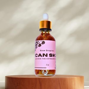 May include: A clear glass bottle of rose-infused serum with a gold cap and dropper, labeled "CAN SKI" and "Rose Droplets." The pink label also reads "ACHIEVE THE EXTRAORDINARY" and "2 oz." The bottle sits on a round, wooden pedestal.