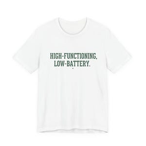 Può includere: T-shirt bianca con la scritta "HIGH-FUNCTIONING, LOW-BATTERY" in verde scuro, carattere serif. La maglietta ha girocollo e maniche corte. Il testo è centrato sul davanti.