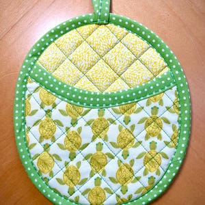 May include: Round quilted potholder with a green polka dot border and hanging loop. The top section features a yellow floral pattern, while the bottom showcases a turtle design in yellow and green on a white background.
