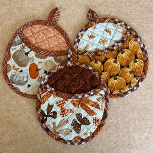 May include: Three round quilted fabric pot holders with hanging loops. One features orange pumpkins, one has white pumpkins with orange accents, and the third displays brown and orange bows. All have plaid trim and stitched patterns.