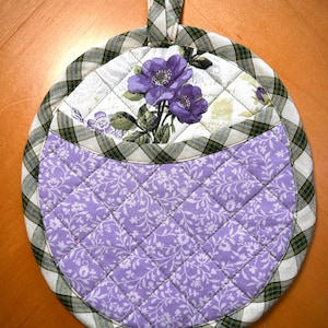 May include: Round quilted pot holder with a loop for hanging. The top section features a white background with purple floral accents. The center is a light purple fabric with a floral pattern. The border is a green and white plaid.