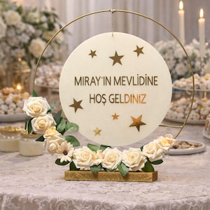 May include: A decorative sign with the text "MIRAY'IN MEVLİDİNE HOŞ GELDINIZ" and star accents. The sign is surrounded by a floral arrangement of cream-colored roses and greenery, set on a gold-colored base and frame.