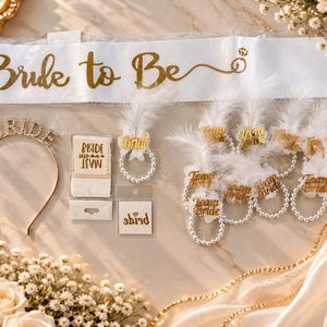 Könnte beinhalten: Eine weiße Satinschärpe mit goldenem Schriftzug Bride to Be, ein goldenes Stirnband mit Strassbuchstaben BRIDE, eine kleine quadratische Karte mit Bride Team Text, eine Karte mit Diamantsymbol und mehrere weiße Feder-Perlen-Armbänder.