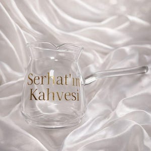 Puede incluir: Una cafetera de cristal transparente con un mango largo, sobre una superficie de satén blanco. Las palabras "Serhat'ın Kahvesi" están impresas en letras doradas en la parte delantera. El fondo es blanco suave y borroso.