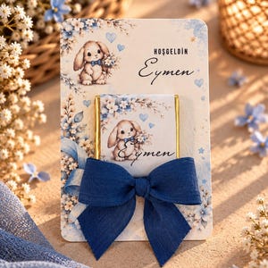 May include: A rectangular card featuring a light blue floral pattern and a cartoon bunny illustration. The text reads Hoşgeldin Eymen. A chocolate bar with matching bunny graphics is attached to the center, adorned with a large, dark blue fabric bow.