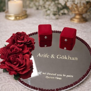 May include: A mirrored tray with a burgundy beaded edge holds two red velvet ring boxes, each with a small diamond. The tray is engraved with names and a quote. A bouquet of red roses and a lit candle are in the background.