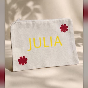 May include: A rectangular canvas pouch with a zip closure. The word "JULIA" is embroidered in yellow, with two red flower accents. The pouch is a neutral colour, with a simple design.