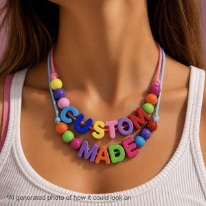 May include: A vibrant necklace spelling out "CUSTOM MADE" in bold, multi-coloured letters. The necklace is strung on a pink and blue cord, interspersed with colourful beads. The necklace is worn around the neck.