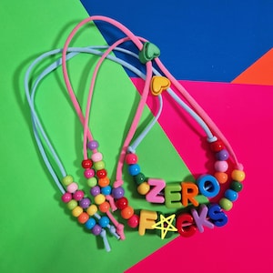 May include: A necklace featuring a pink cord and a light blue cord, adorned with multicoloured plastic beads and heart-shaped toggles. The necklace displays the text ZERO FUCKS in large, colourful block letters, with a yellow star drawn on the letter F.