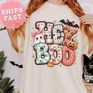 May include: An ivory short-sleeve t-shirt featuring a large graphic print with the text HEY BOO. The design includes a ghost with a pink bow, a spider web, a bat, a jack-o'-lantern, and candy corn, all rendered in a colorful, patterned style.