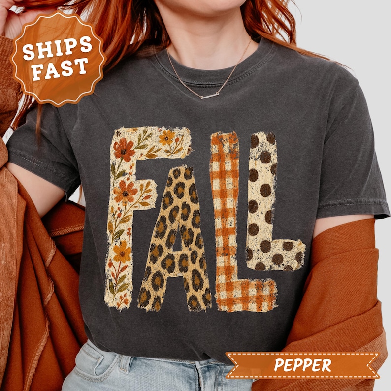 May include: A charcoal gray short-sleeve t-shirt featuring the word FALL printed in large block letters. Each letter has a unique pattern: the F has a floral print, the A has a leopard print, the L has an orange gingham, and the L has polka dots.