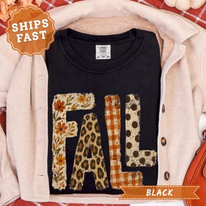 May include: A black short-sleeve t-shirt featuring the word FALL in large block letters. Each letter has a distinct pattern: the F has a floral print, the A has a leopard print, the L has an orange gingham check, and the L has brown polka dots.