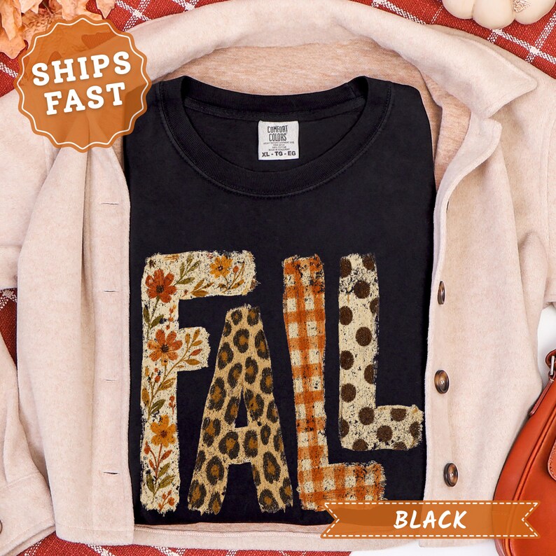 May include: A black short-sleeve t-shirt featuring the word FALL in large block letters. Each letter has a distinct pattern: the F has a floral print, the A has a leopard print, the L has an orange gingham check, and the L has brown polka dots.