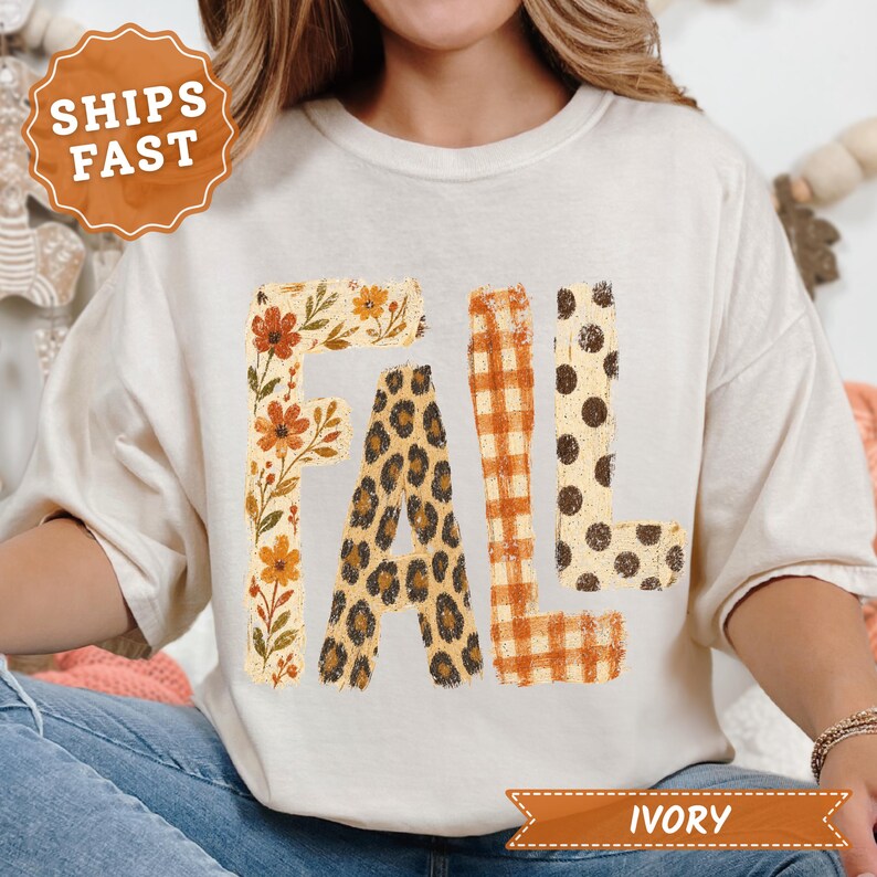 May include: An ivory t-shirt featuring the word FALL printed in large letters. Each letter has a distinct pattern: the F has orange and yellow flowers, the A has a leopard print, the L has an orange plaid, and the second L has brown polka dots.