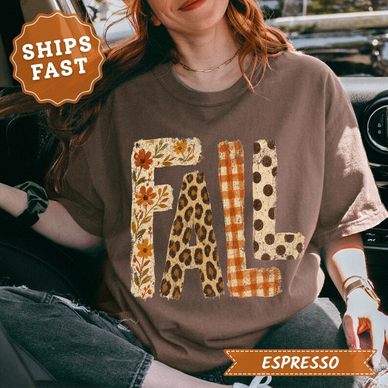 May include: A brown short-sleeve t-shirt featuring the word FALL printed in large block letters. Each letter has a distinct pattern: the F has a floral print, the A has a leopard print, the L has an orange plaid print, and the L has polka dots.