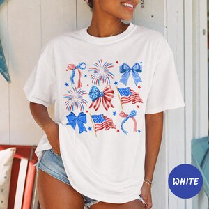 May include: A white short-sleeved t-shirt featuring a graphic print of red, white, and blue patriotic elements including fireworks, American flags, and various bows. The design is arranged in a grid-like pattern across the center of the shirt.