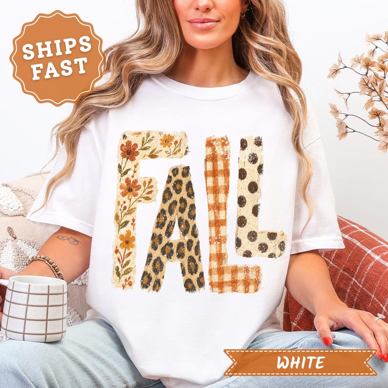 May include: A white short-sleeve t-shirt featuring the word FALL printed in large block letters. Each letter has a unique pattern: the F is floral, the A is leopard print, the L is orange plaid, and the second L has brown polka dots.