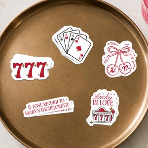 May include: A collection of five vinyl stickers featuring red and white designs including a 777 slot machine symbol, a hand of playing cards, cherries with a bow, a slot machine with the text Lucky in Love, and a label reading If lost, return to Mary's bachelorette.