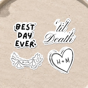 May include: Four white stickers with black ink designs. One reads BEST DAY EVER, another reads til Death, one features a banner with H+M and two birds, and the last is a heart shape with H+M inside. The stickers are arranged on a round tray.