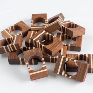May include: A collection of square wooden napkin rings. Each ring is made of dark brown wood with inlaid light-colored wood stripes. The rings have a circular opening in the center. The napkin rings are scattered on a white surface.