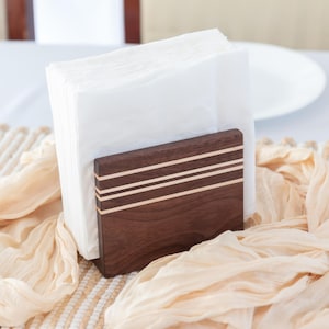 May include: A wooden napkin holder with a dark brown finish and three horizontal light-colored stripes. The holder is filled with white napkins and sits on a table with a cream-colored runner and a white tablecloth. A candle and tableware are also visible.