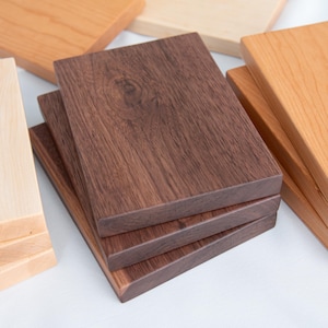 May include: A collection of wooden coasters in various wood tones, including light, medium, and dark brown. The coasters are square and stacked in groups, showcasing the natural wood grain.