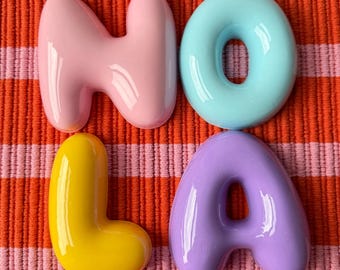Puede incluir: Cuatro letras tridimensionales brillantes que forman la palabra NOLA. La letra N es rosa claro, la O es azul claro, la L es amarillo brillante y la A es violeta claro. Cada letra tiene una forma redondeada y lisa, similar a una burbuja.
